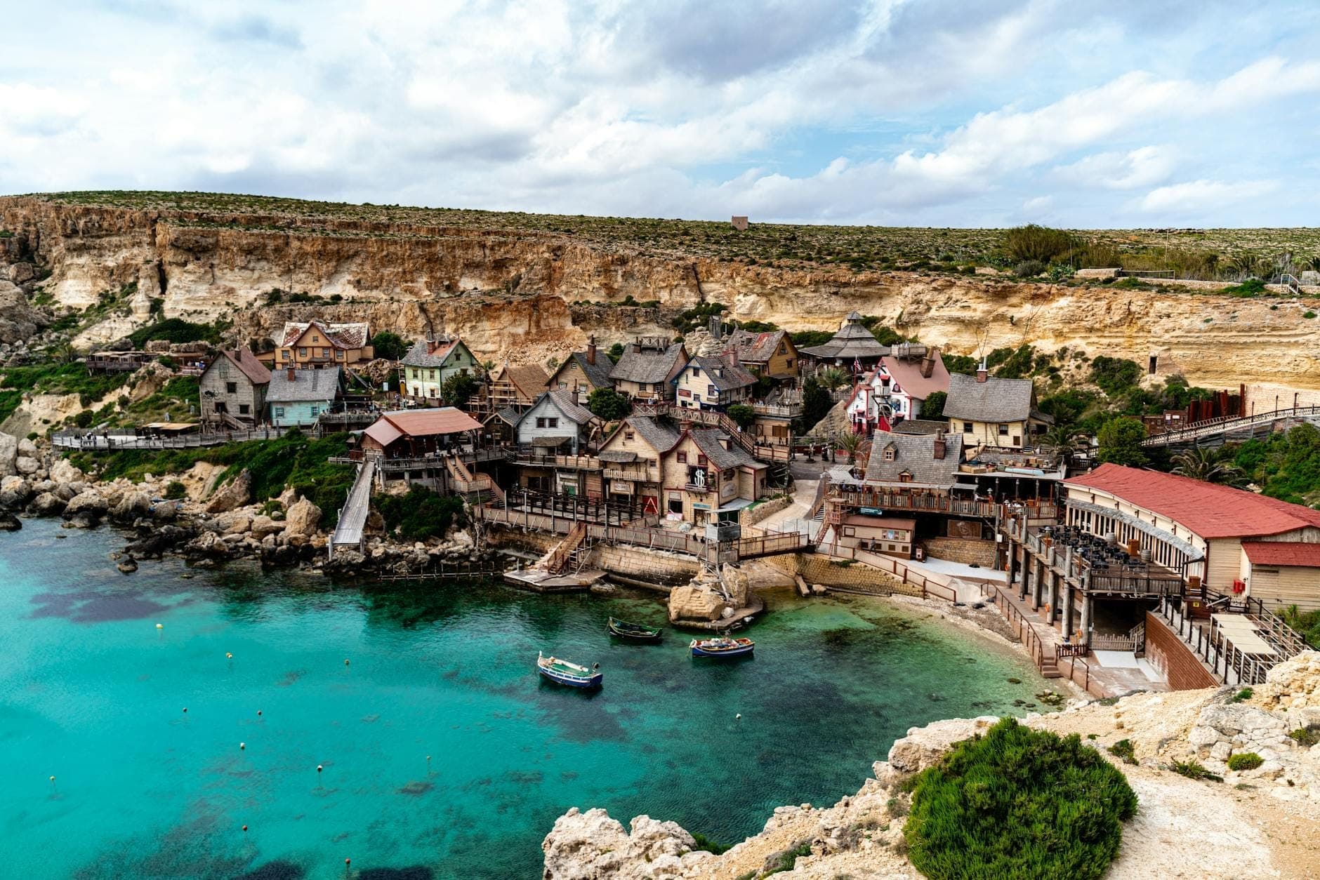 Ein weiter, farbenfroher Blick auf die schrägen Holzhäuser des Popeye Village, eingebettet an der türkisblauen Anchor Bay, mit Klippen im Hintergrund unter strahlend blauem Himmel.