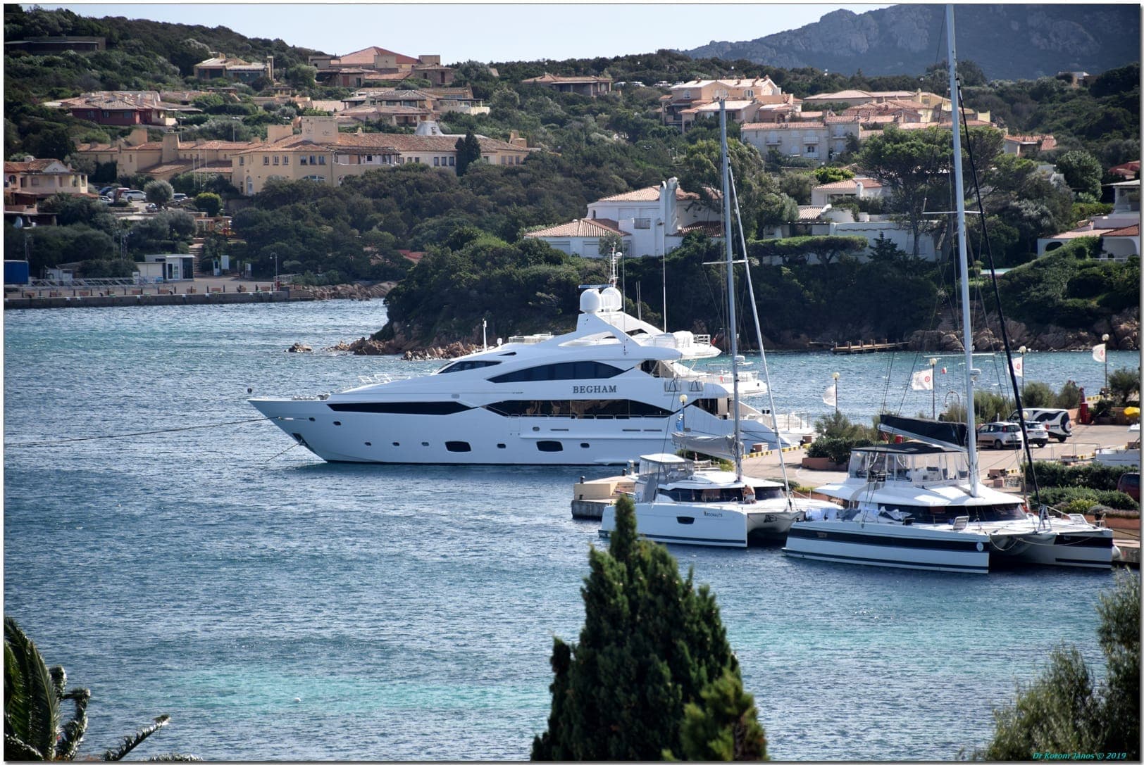 Gran superyate blanco y catamaranes atracados en el Puerto Cervo Marina, con villas en la ladera y exuberante vegetación al fondo.