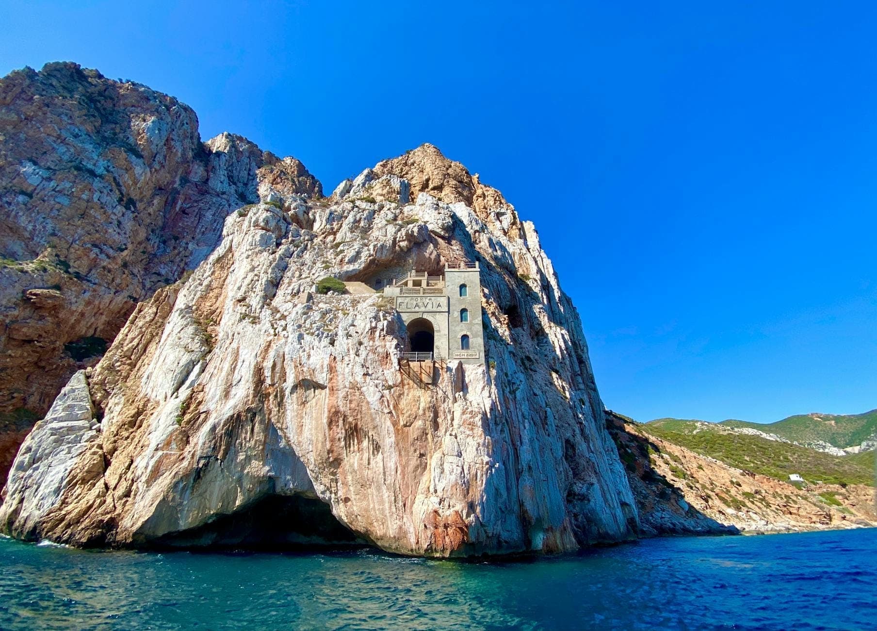 Porto Flavia, el puerto minero en los acantilados, encajado dramáticamente en blancas paredes de caliza sobre un mar azul, bajo un cielo despejado, con la costa de Cerdeña al fondo.