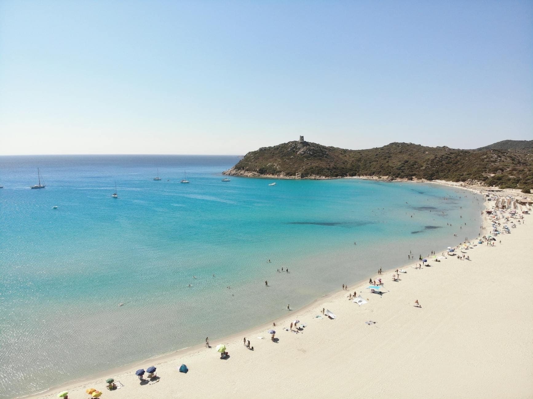 Luftaufnahme des Strandes Porto Giunco mit türkisblauem Wasser, feinem weißen Sand, Menschen beim Entspannen und dem spanischen Wachturm auf einem Hügel im Hintergrund.