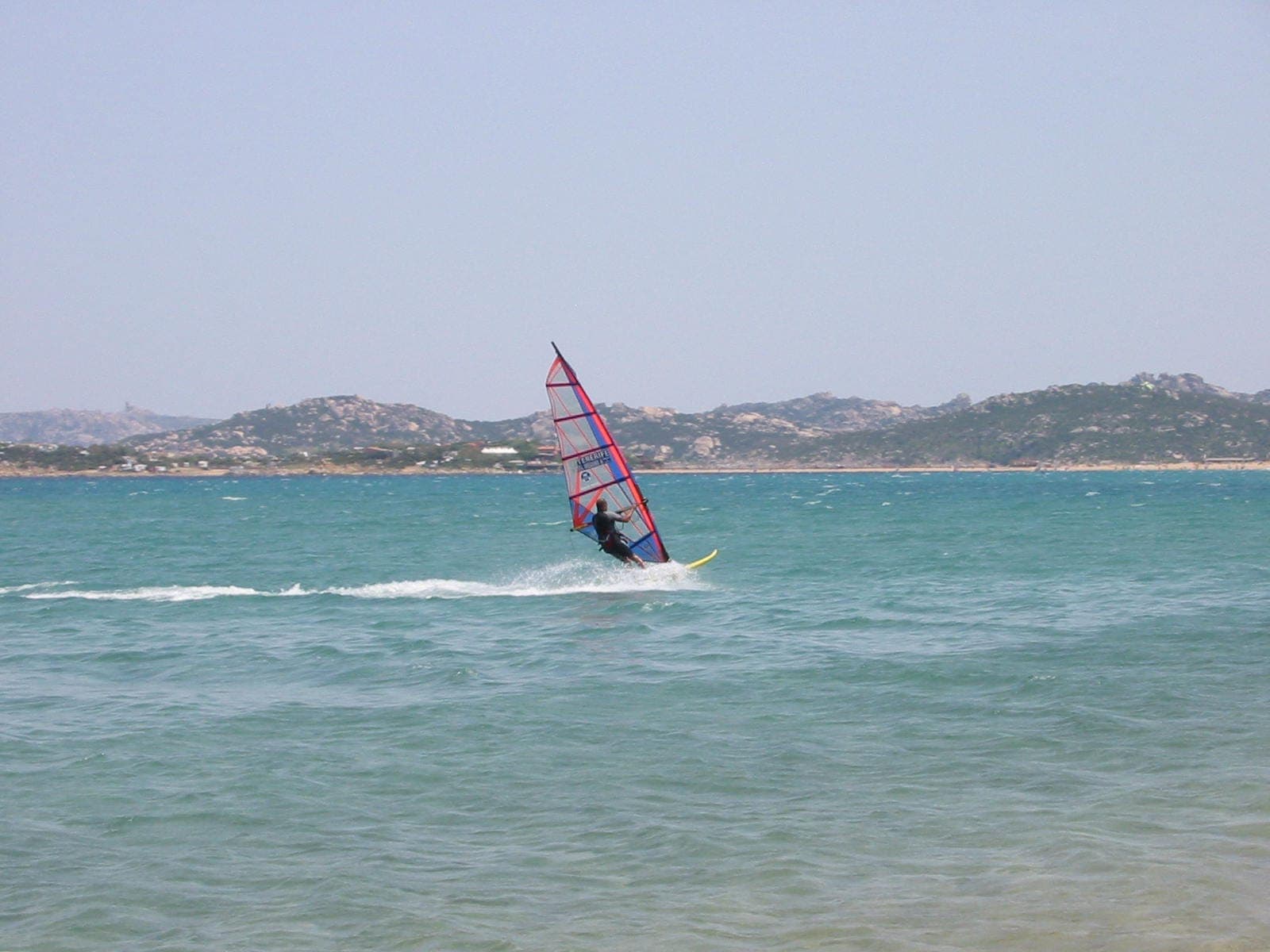 Un windsurfista se desliza sobre las aguas turquesas de Porto Pollo, con la rocosa costa de Cerdeña visible bajo un cielo azul despejado.