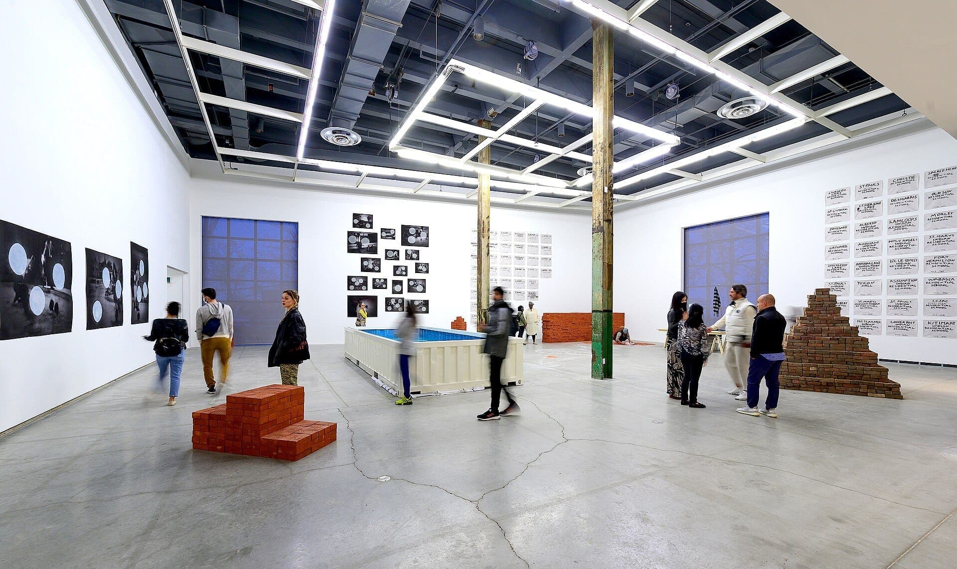 Interno della The Power Plant Contemporary Art Gallery con visitatori che osservano installazioni moderne, fotografie su pareti bianche e travi industriali a vista sotto un'illuminazione intensa.