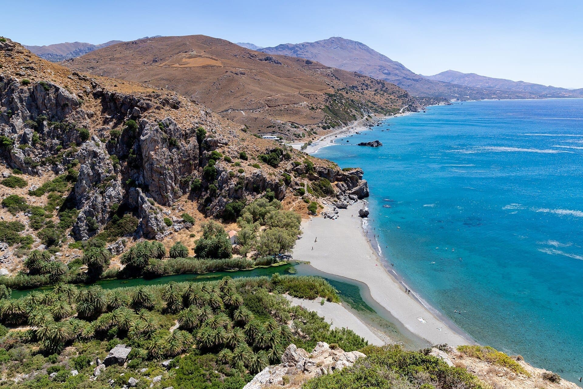 Panoramablick auf den Preveli Beach auf Kreta mit einem Palmenhain, türkisfarbenem Wasser, Sandstrand und schroffen Bergen.