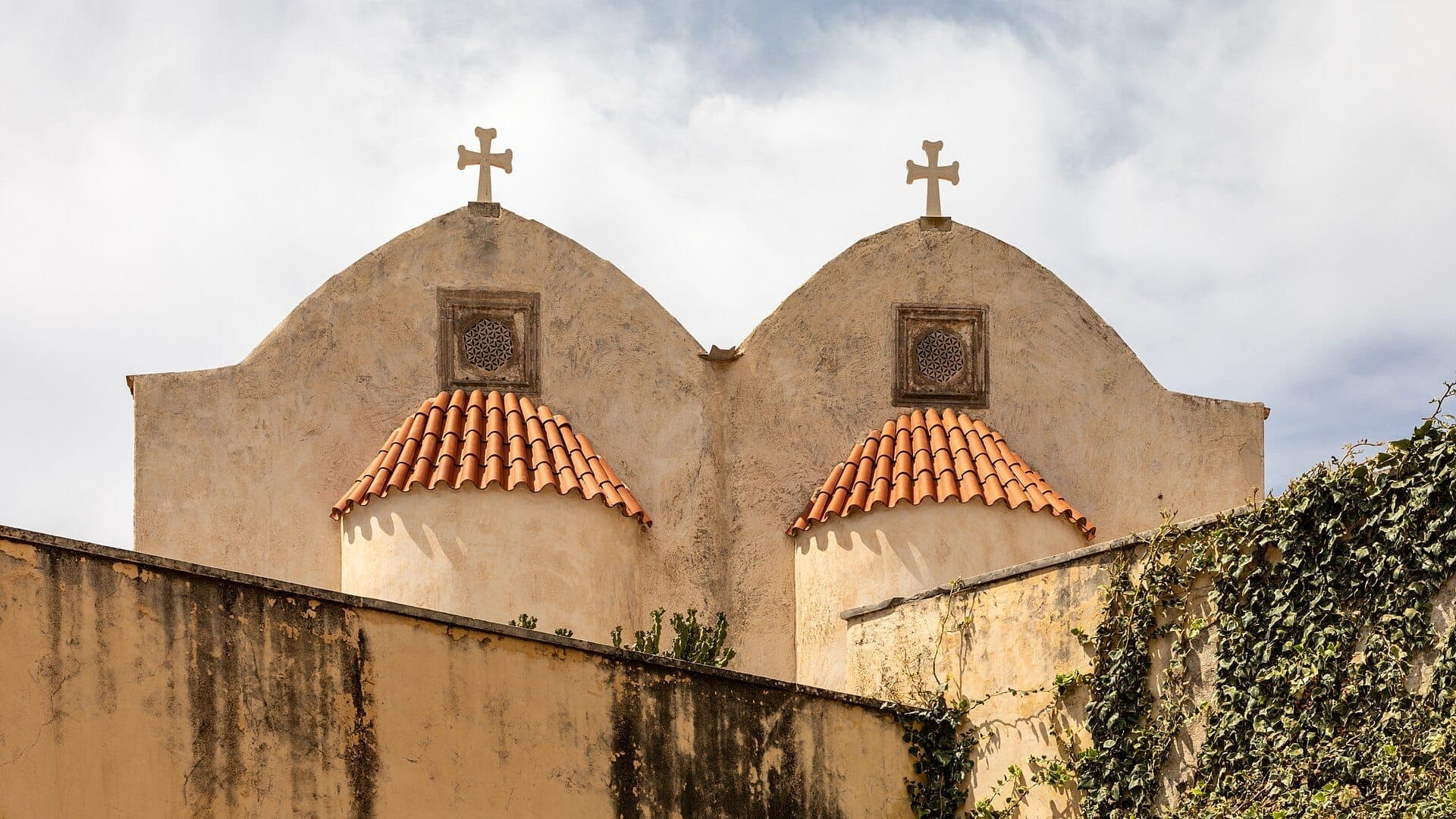 Zwei Kuppeln mit roten Ziegeldächern und Kreuzen auf dem Moni Preveli Kloster, eingerahmt von verwitterten Mauern und kriechenden Efeu unter einem hellen Himmel.