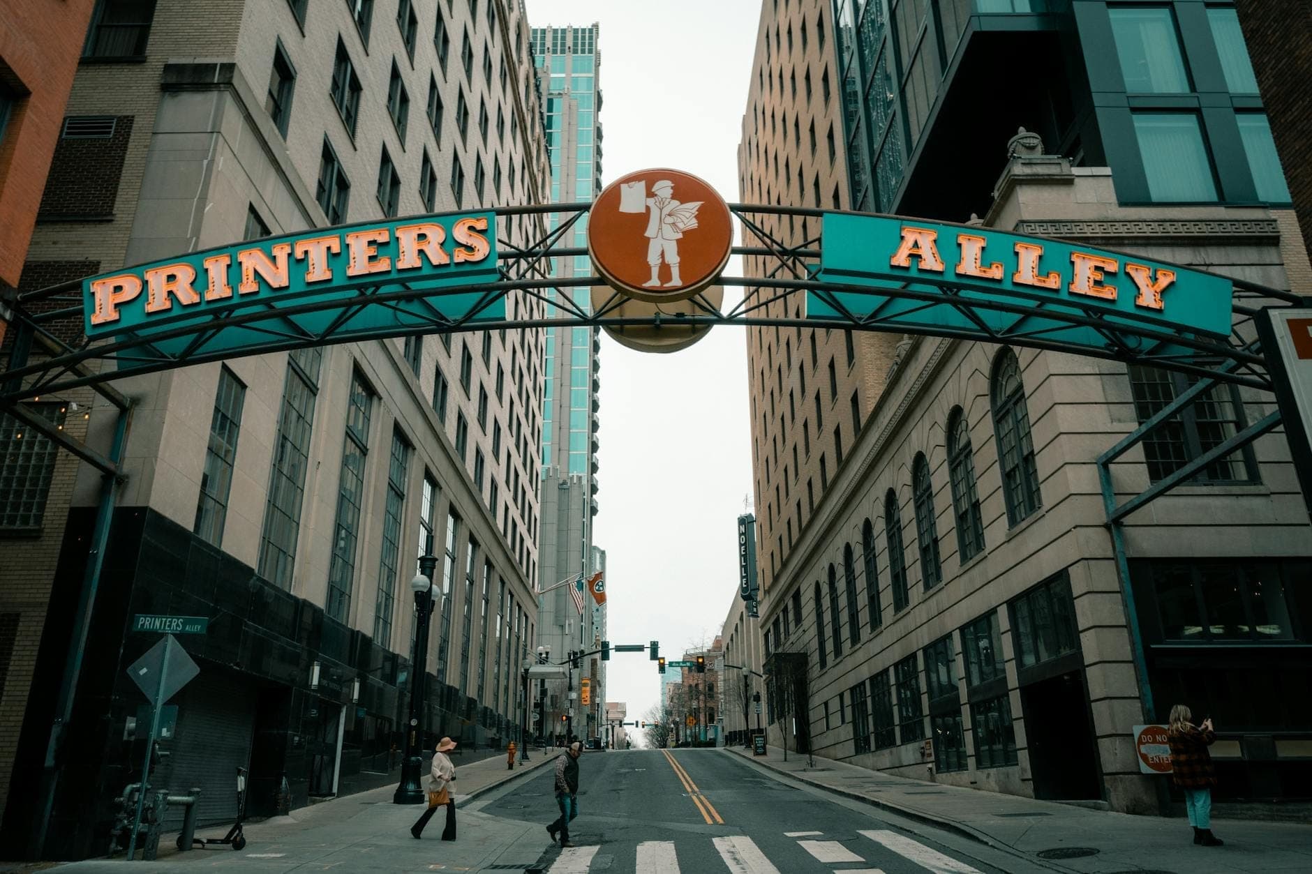 The iconic Printer's Alley sign arches over a quiet downtown Nashville street, flanked by tall historic buildings and a few people walking.