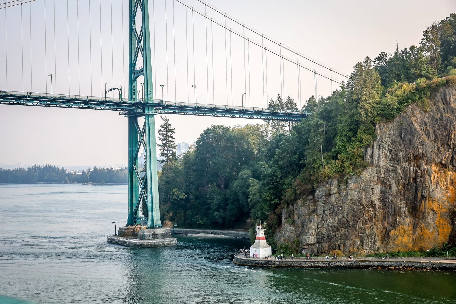 วิวของ Lions Gate Bridge จาก Prospect Point โดยมีเสาสะพาน หน้าผาที่ปกคลุมด้วยป่า และประภาคารขนาดเล็กตามแนวทางเดินริมน้ำของ Stanley Park