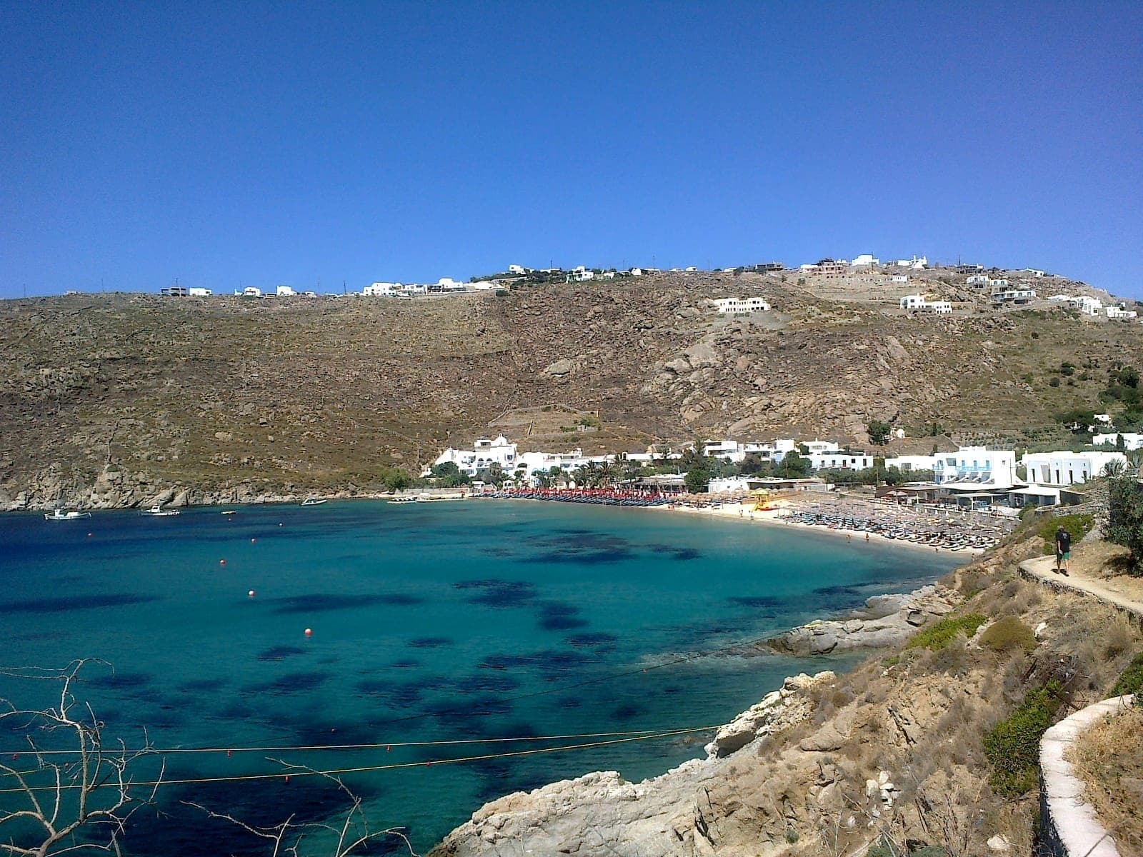 Psarou Beach auf Mykonos mit türkisfarbenem Wasser, hellem Sand, Liegestühlen und weißen Gebäuden vor einem felsigen Hang unter klarem blauem Himmel.