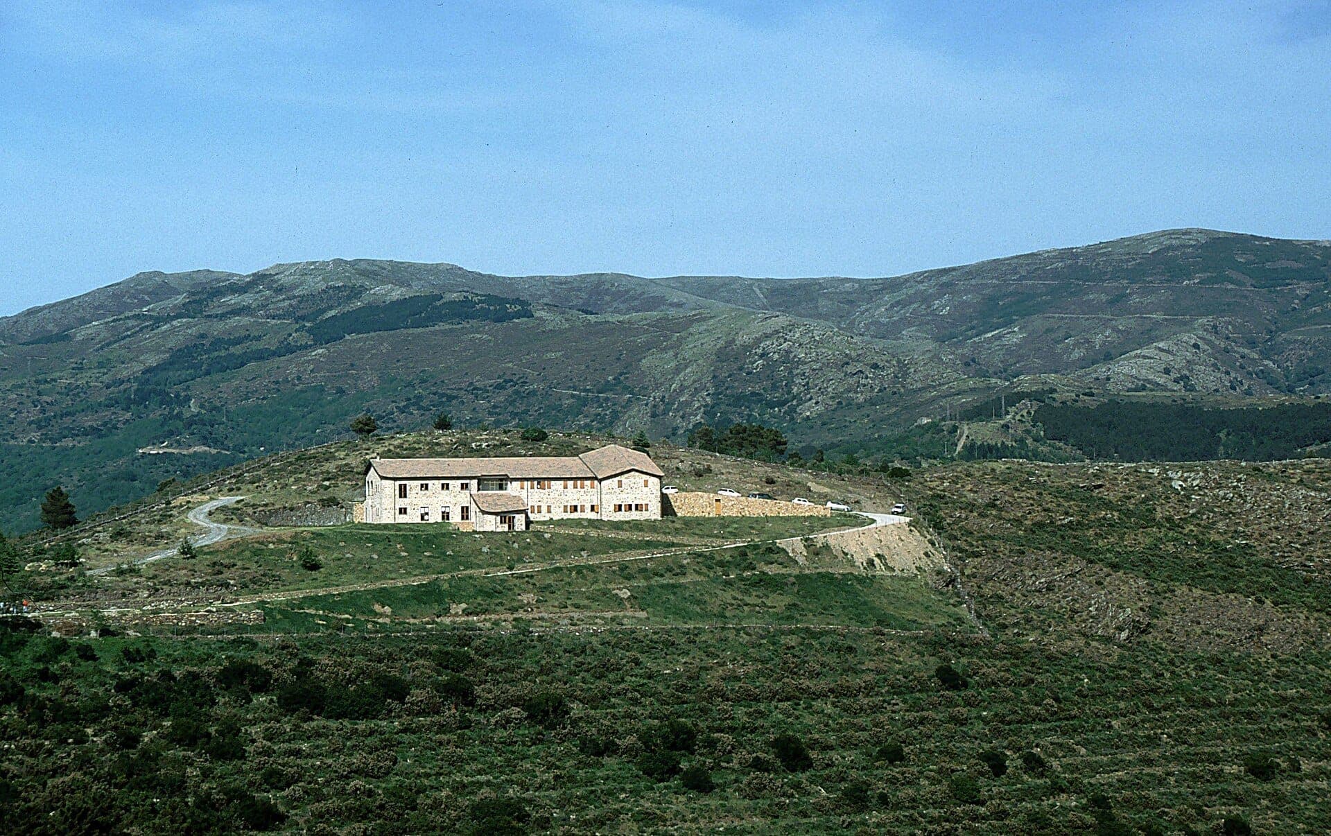 Albergue de piedra rodeado de caminos sinuosos y escarpadas colinas verdes en Punta La Marmora, con las montañas del Gennargentu al fondo.