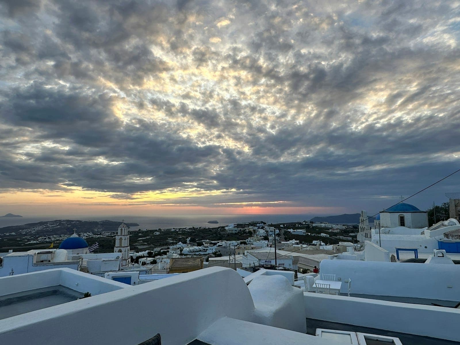 Malerischer Blick über das Dorf Pyrgos bei Sonnenuntergang mit weißgetünchten Häusern, blauen Kuppeln und dramatisch bewölktem Himmel – die friedliche Hügellage im Zentrum Santorinis.