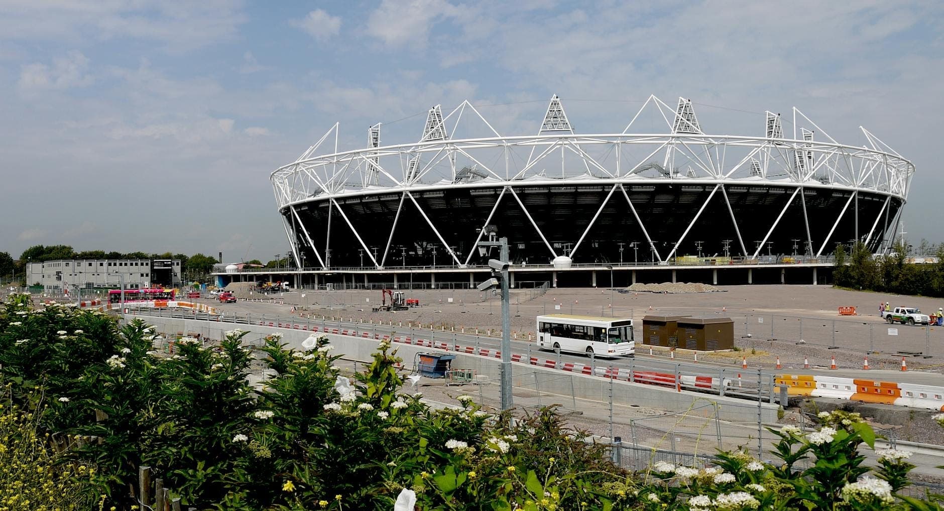 Weitwinkelaufnahme des Londoner Olympiastadions, umgeben von offenen Flächen, Grünflächen und Absperrungen an einem hellen Tag im Queen Elizabeth Olympic Park.