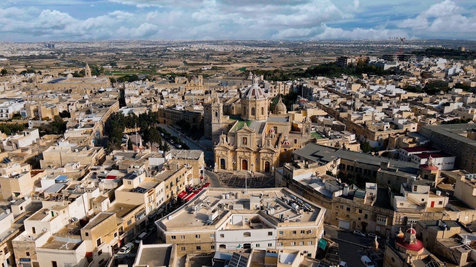 Luftaufnahme von Rabat, Malta, mit einer historischen Kirche im Zentrum, umgeben von Kalksteingebäuden und weiter Landschaft unter einem teils bewölkten Himmel.