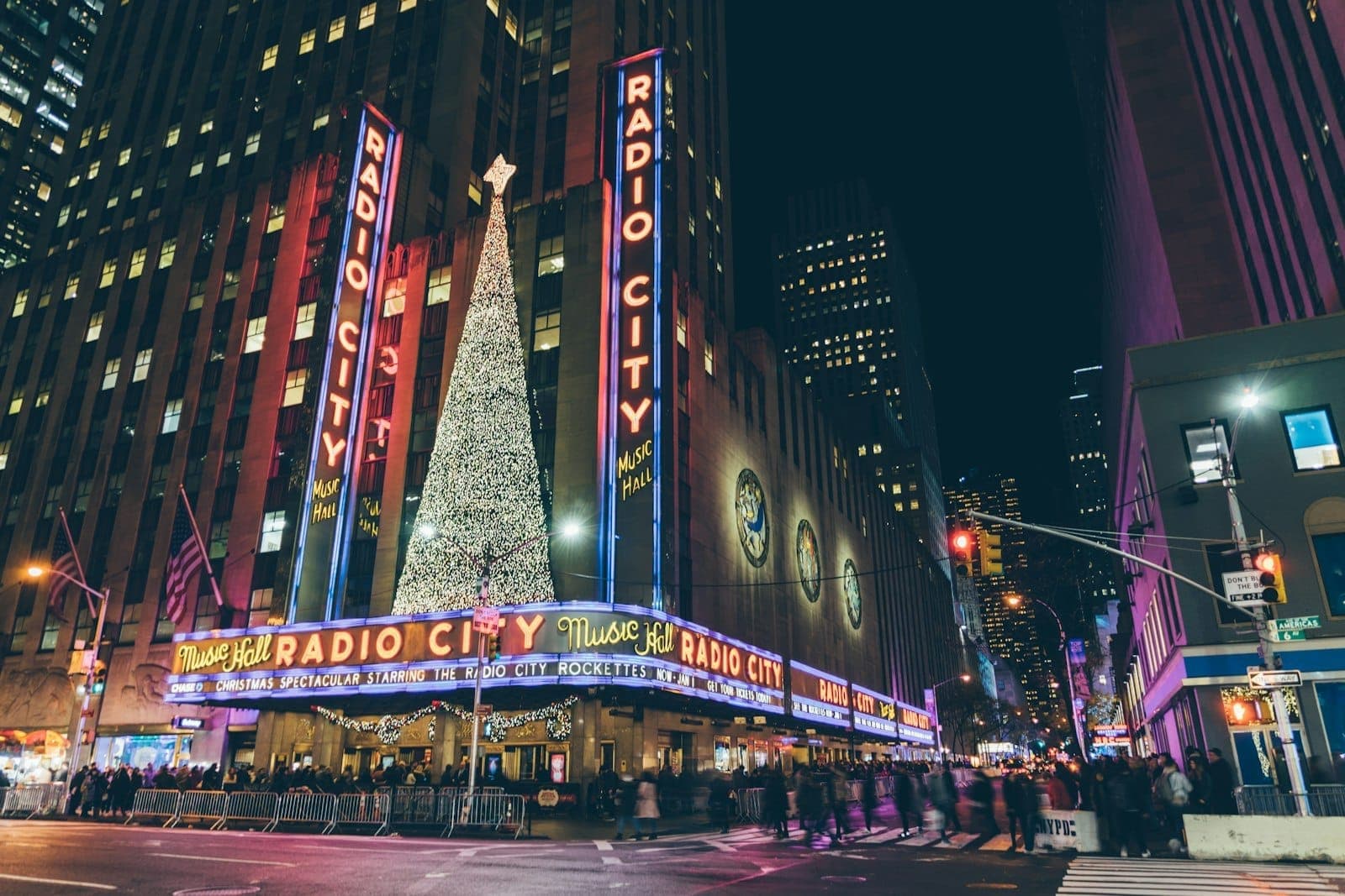 วิวกลางคืนของ Radio City Music Hall ใจกลางแมนฮัตตัน พร้อมไฟนีออนสุดไอคอนิค ป้าย Art Deco และต้นคริสต์มาสประดับไฟขนาดใหญ่หน้าทางเข้า