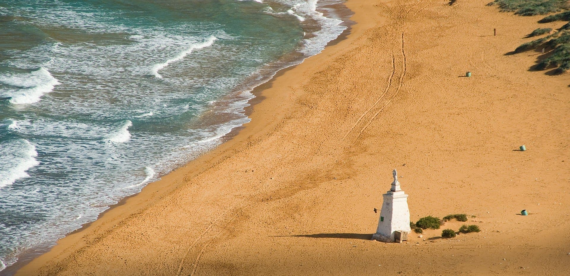 Luftaufnahme der roten Sandküste der Ramla Bay, sanfte blaue Wellen, ein einzelnes Statuen-Monument und vereinzelte Begrünung entlang des breiten Strandes.