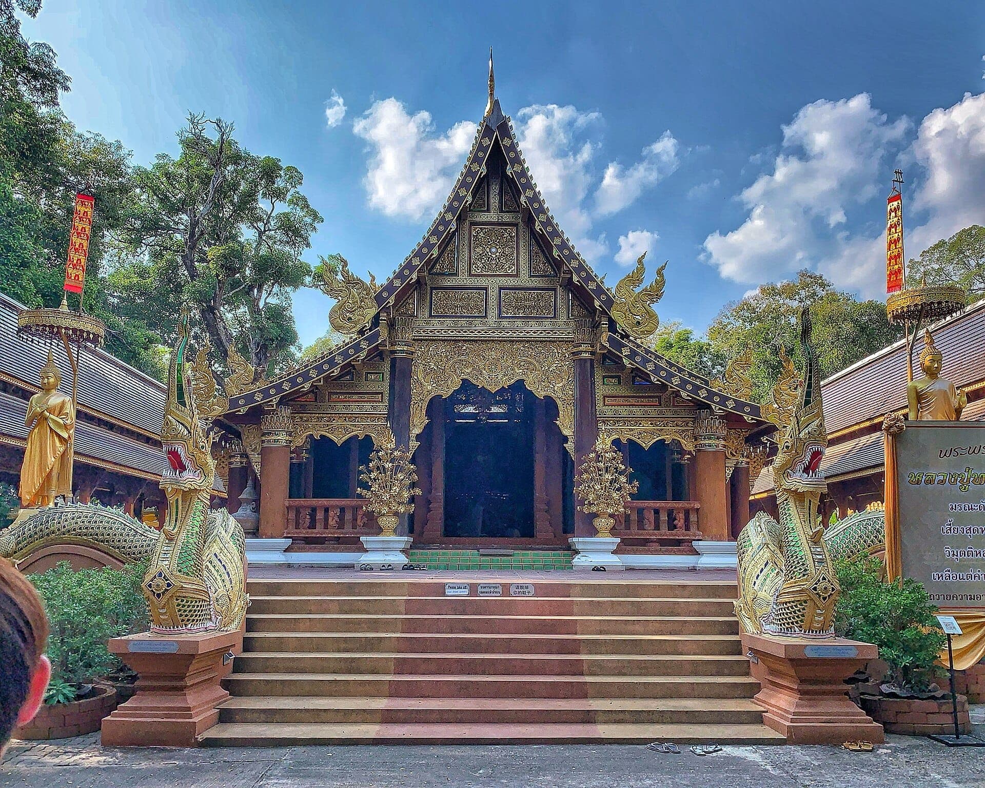 Verzierter Holztempel im Wat Ram Poeng mit goldenen Statuen, Naga-Schlangen-Geländern, üppigen Bäumen und blauem Himmel in Chiang Mai, Thailand.