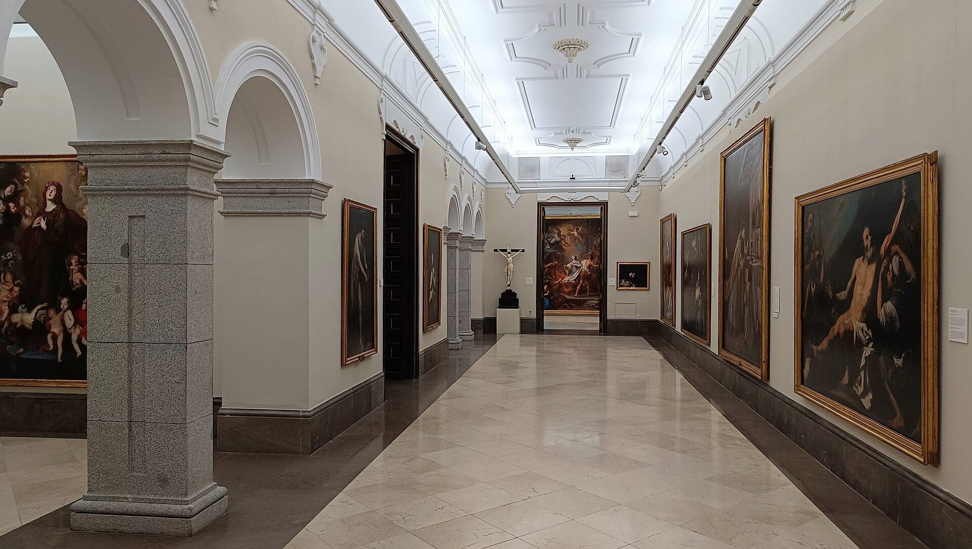 Weitläufige Galerie mit Marmorboden in der Real Academia de Bellas Artes de San Fernando, gesäumt von großen gerahmten Gemälden und klassischen Säulen unter hellem Licht.