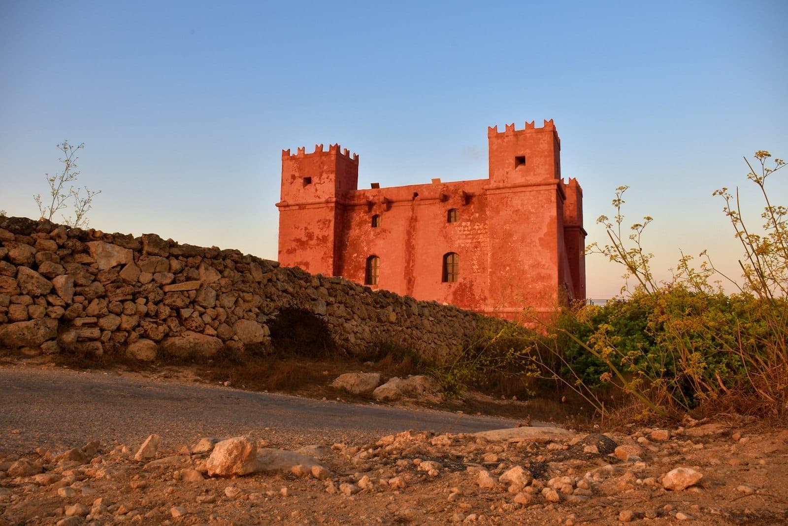 Der Red Tower auf Malta erhebt sich über einer Trockensteinmauer, seine rote Fassade im warmen Abendlicht vor einem klaren blauen Himmel.
