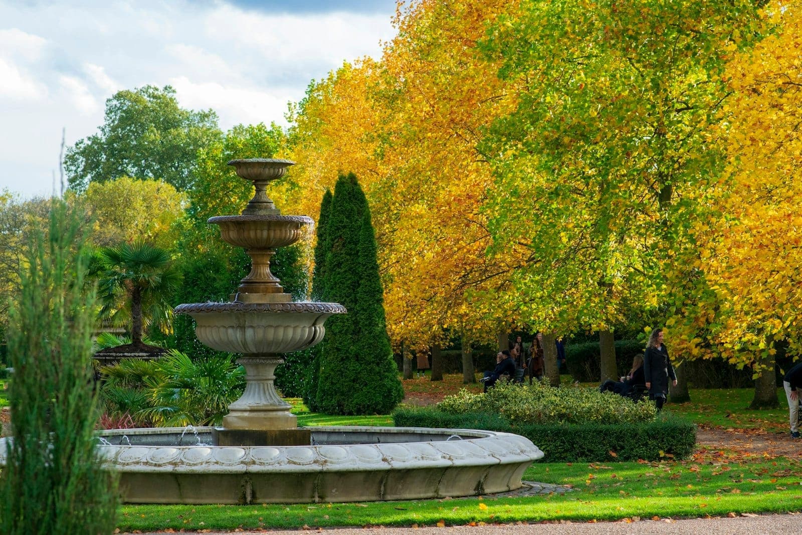 Centralna kamienna fontanna otoczona bujnymi trawnikami i złotymi jesiennymi drzewami w Regent's Park, ze spacerującymi i odpoczywającymi na ławkach odwiedzającymi.
