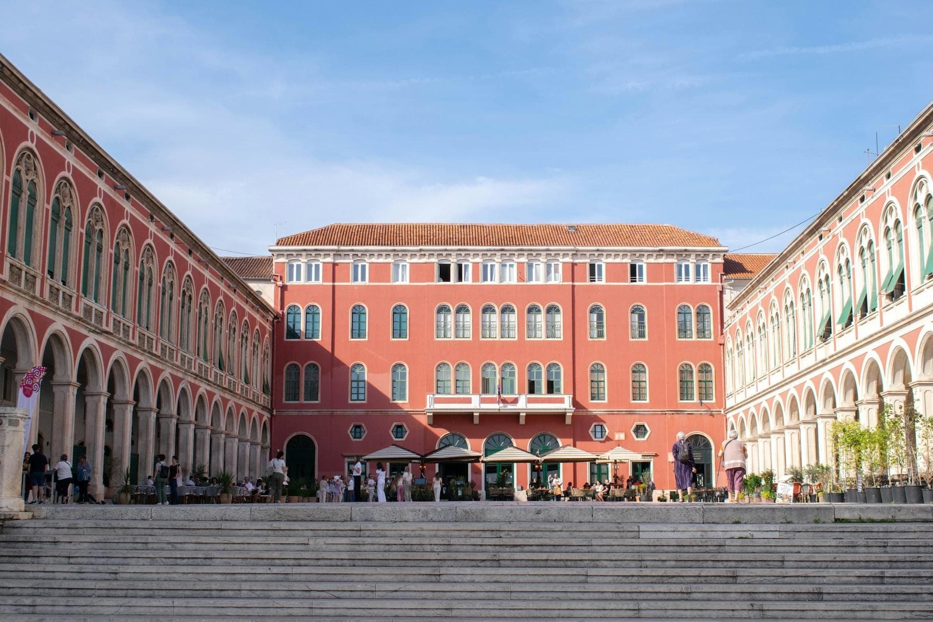 Vista panoramica della Piazza della Repubblica (Prokurative) a Spalato, Croazia, con le facciate neorinsasimentali rosse, i portici ad arco e le persone che si godono la piazza aperta sotto un cielo azzurro.