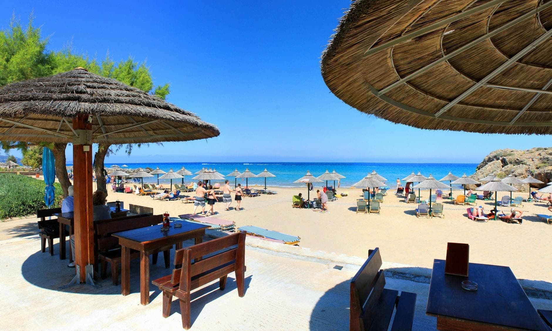 Blick von einer Strandtaverne am Rethymno Beach mit Strohsonnenschirmen, Liegestühlen, entspannenden Menschen und klarem blauem Meer unter einem sonnigen Himmel.