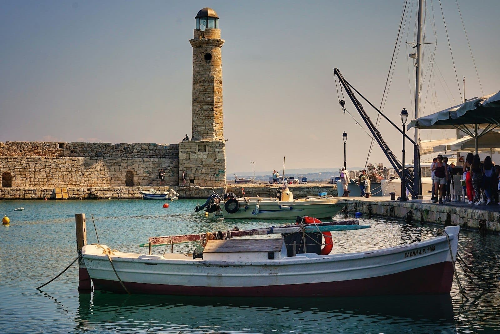 Leuchtturm und Fischerboote im historischen venezianischen Hafen von Rethymno, Spaziergänger am Ufer, ruhiges türkisfarbenes Wasser und alte Steinarchitektur im warmen Nachmittagslicht.