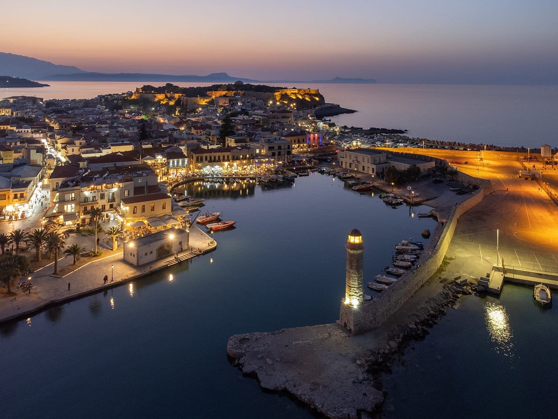 Luftaufnahme der Altstadt von Rethymno in der Abenddämmerung mit beleuchtetem venezianischem Hafen, steinigem Leuchtturm, Cafés an der Uferpromenade und mittelalterlicher Festung in der Ferne.