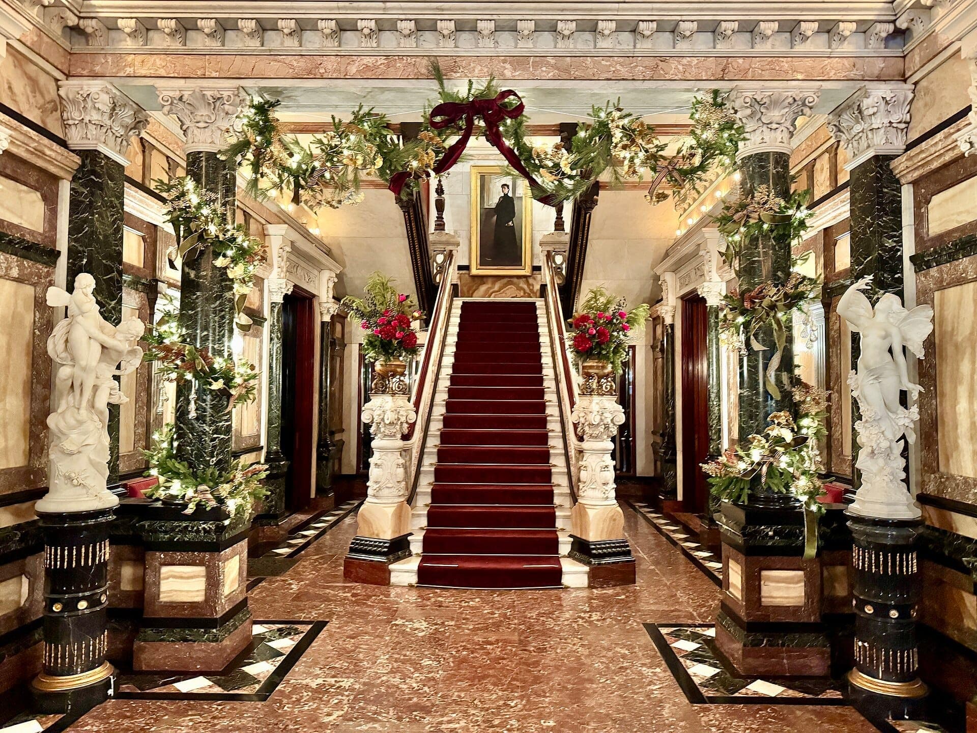 Grand entryway of the Richard H. Driehaus Museum featuring marble floors, ornate columns, festive greenery, and a sweeping red-carpet staircase.