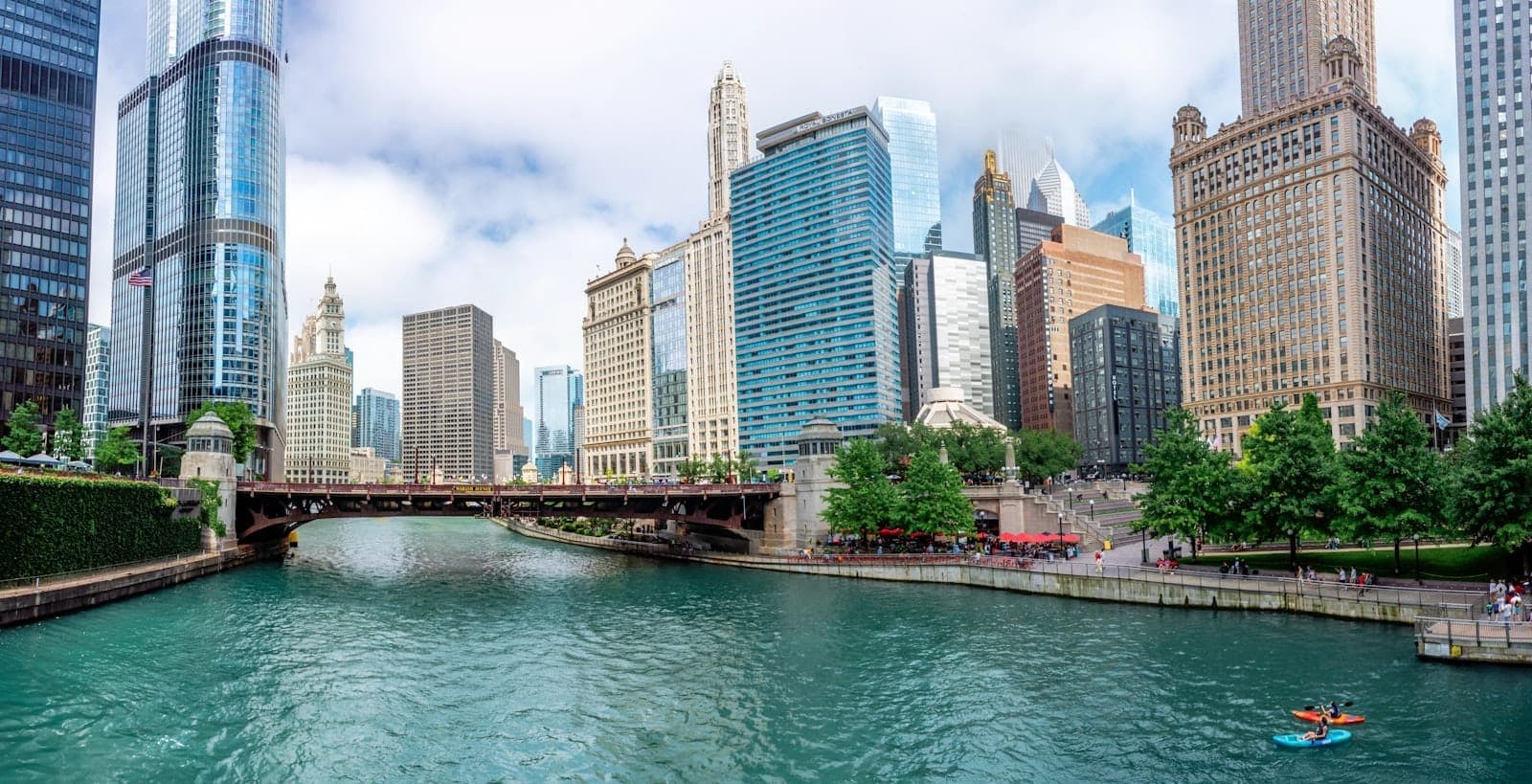 Wide view of the Chicago River in River North with iconic skyscrapers, a bridge, lush greenery, and a kayaker on the water under a bright sky.