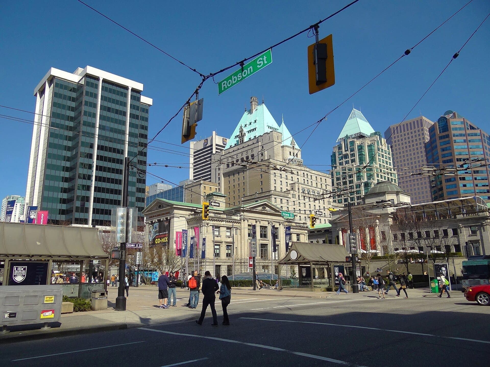 วิวของ Robson Street ในย่านดาวน์ทาวน์ Vancouver พร้อมตึกระฟ้าสมัยใหม่ อาคารประวัติศาสตร์ และผู้คนกำลังข้ามถนนใต้ท้องฟ้าสีฟ้าใส