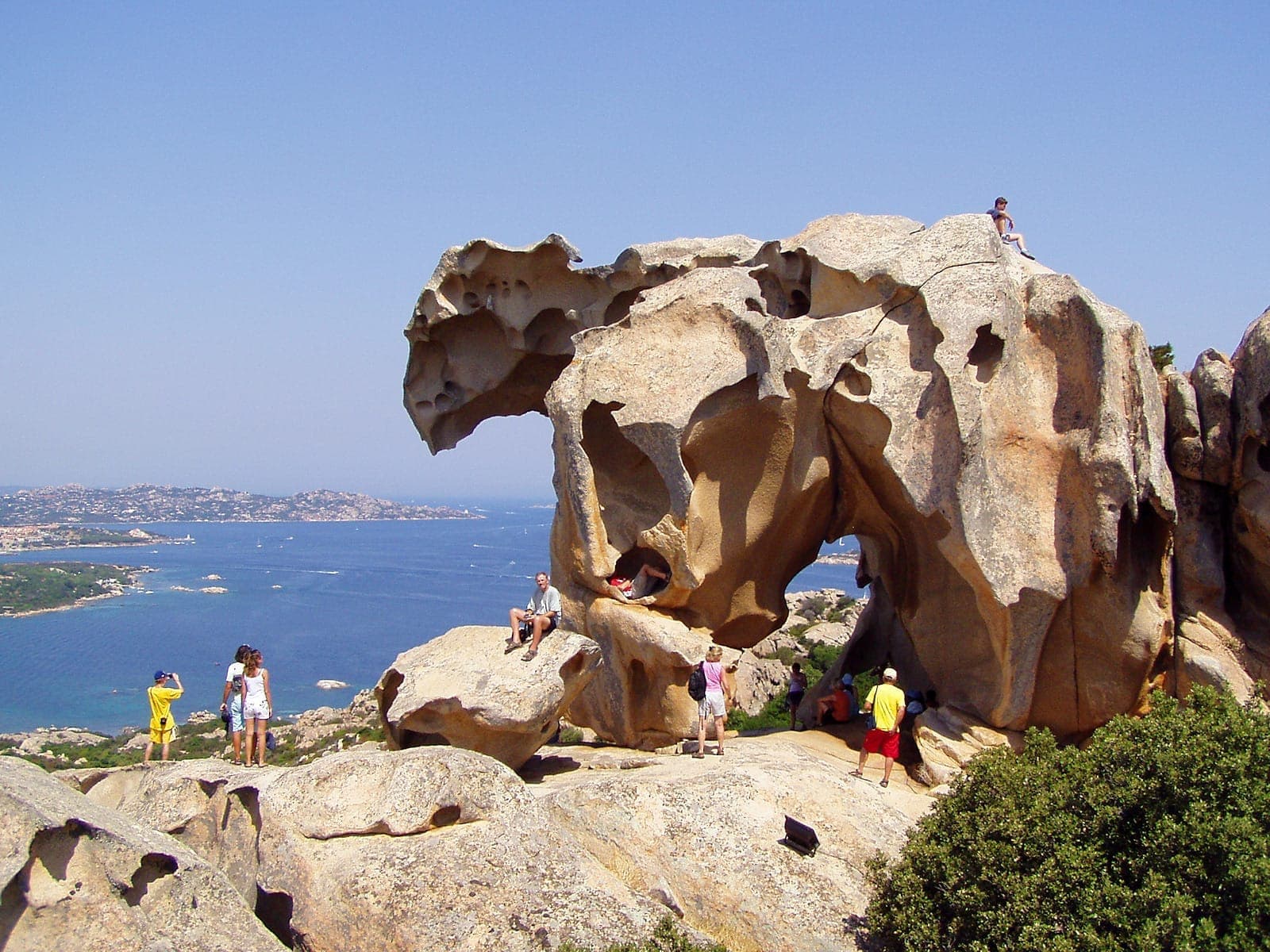 Turistas explorando la Roccia dell'Orso, una formación de roca granítica tallada por el viento con vistas a la costa azul y las exuberantes colinas verdes de Palau, Cerdeña.