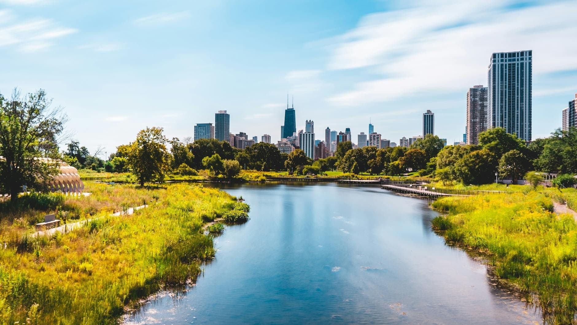 A view of Chicago’s skyline from Lincoln Park, showing a calm pond, walking paths, and lush greenery on a sunny day.