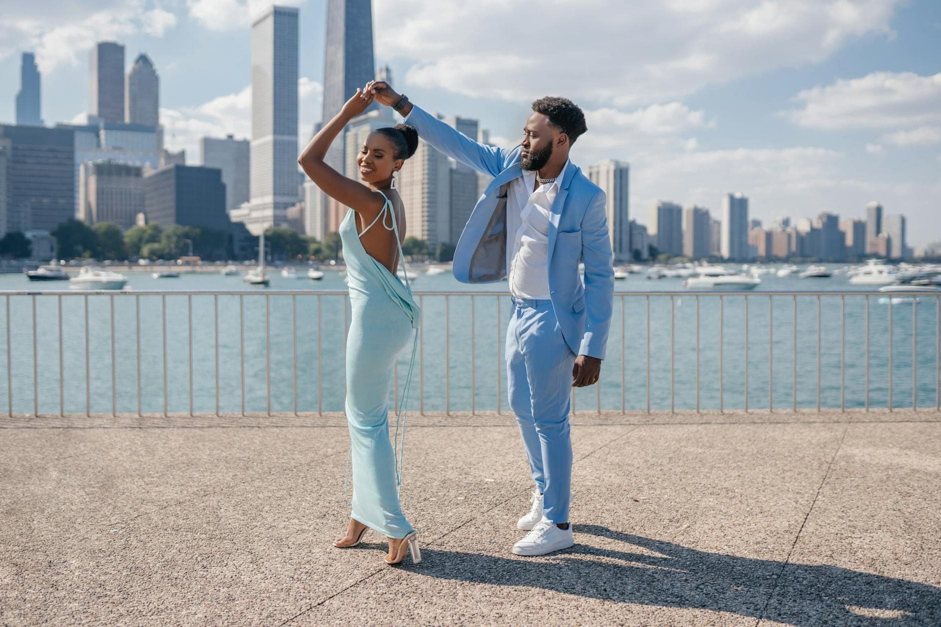 A stylish couple dances together on a waterfront promenade with the Chicago skyline and boats in the background on a sunny day.