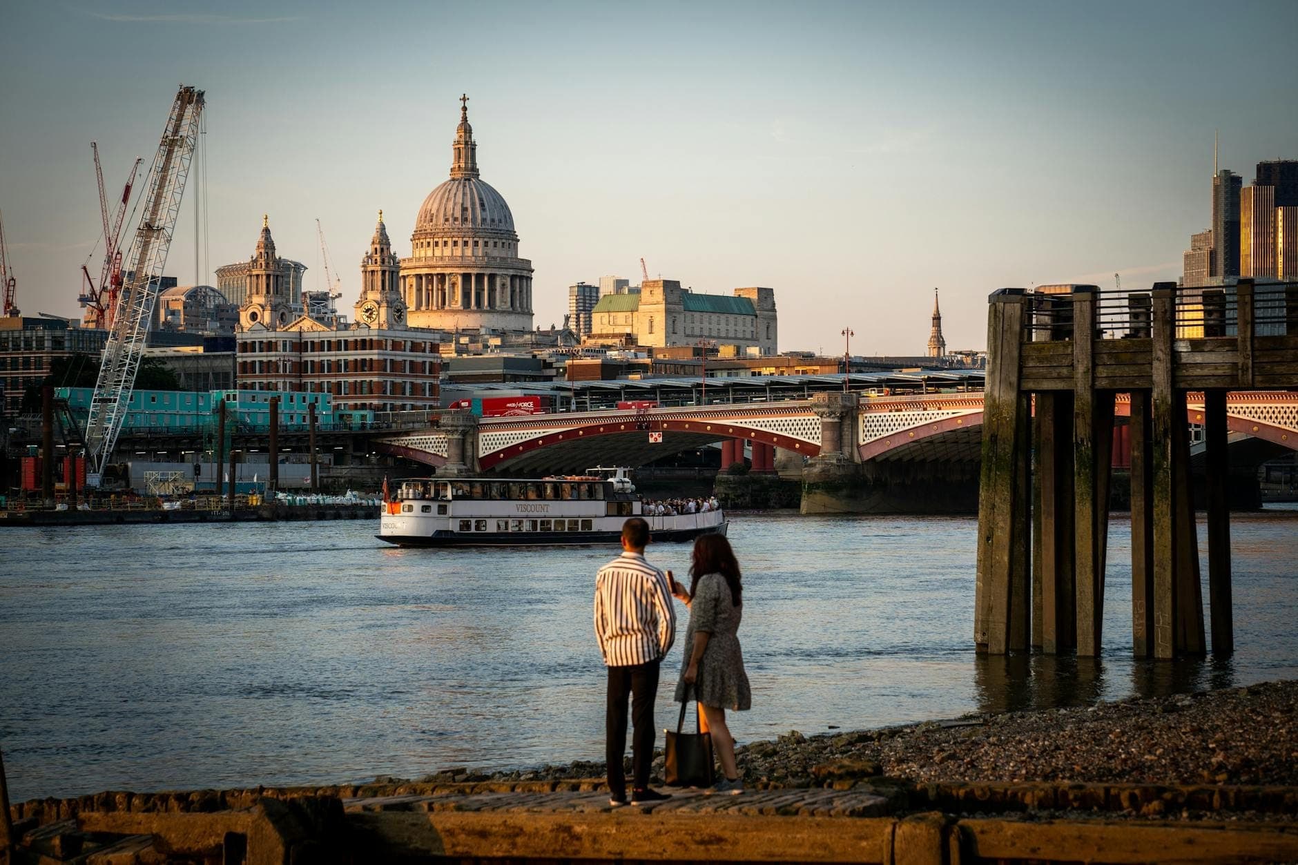 Un couple se tient au bord de la Tamise au coucher du soleil, regardant vers la cathédrale Saint-Paul et un bateau qui passe, avec la skyline de Londres en arrière-plan.