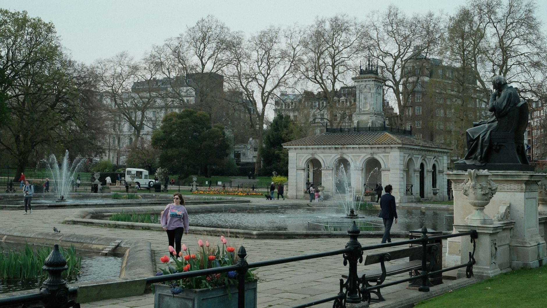 Des personnes se promènent dans les Italian Gardens à Hyde Park, avec un historique pavillon de pompage, des fontaines, des statues, des tulipes et l'architecture de Londres en arrière-plan.