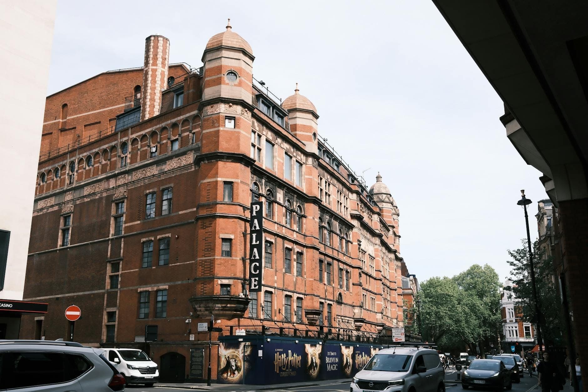 Vue de la rue du célèbre Palace Theatre de Londres avec sa façade en briques rouges, ses enseignes visibles et des voitures garées à l'extérieur en journée.