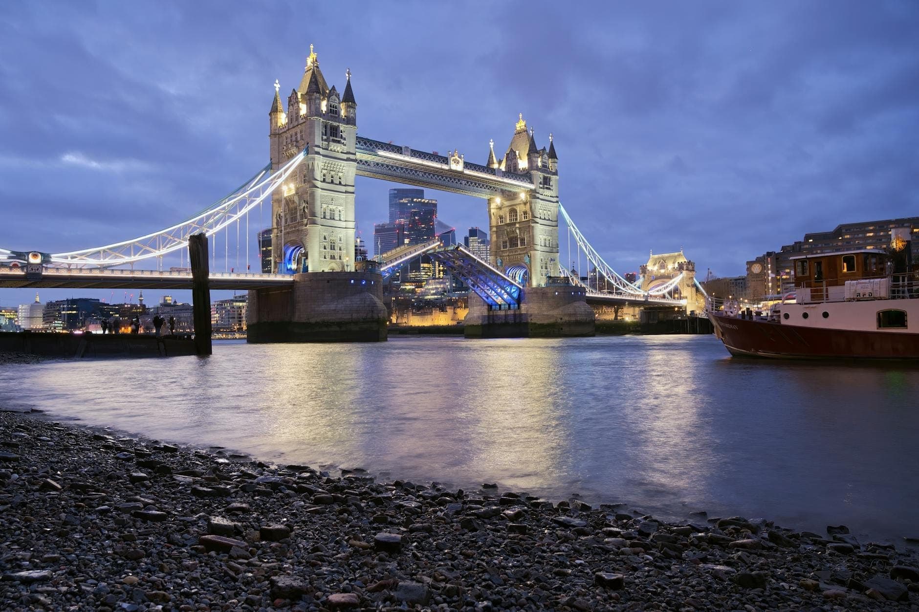 Tower Bridge illuminé au crépuscule avec les lumières de la ville se reflétant sur la Tamise et un bateau à proximité, capturant une soirée romantique à Londres.