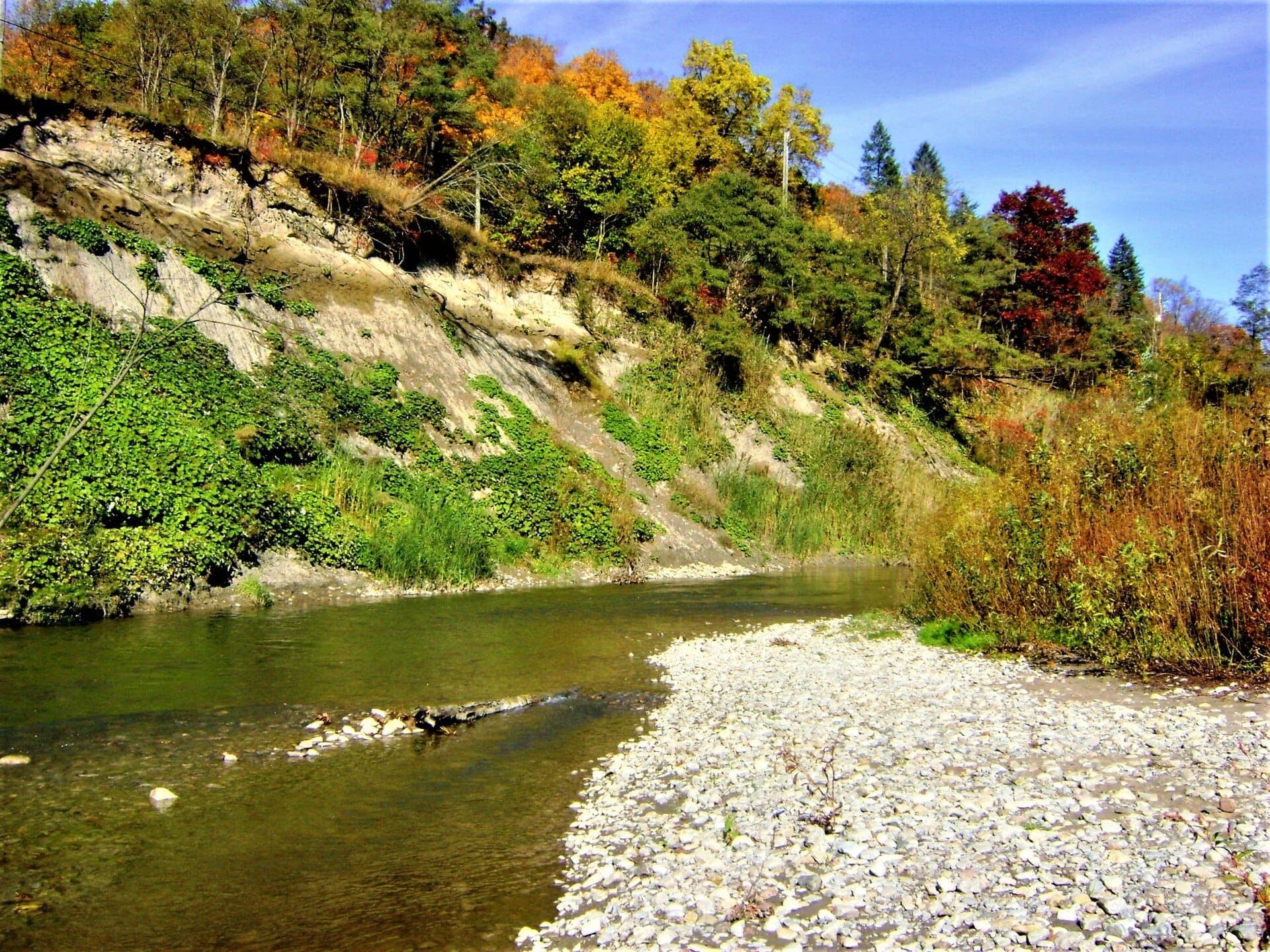 Arroyo poco profundo serpenteando por una orilla rocosa y colinas boscosas en Rouge National Urban Park bajo un brillante cielo azul de otoño.