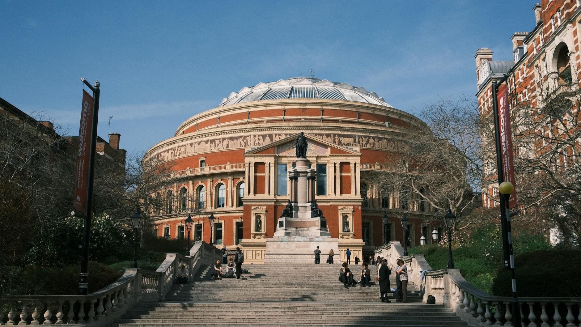 Vista panorámica del Royal Albert Hall en Londres, con su icónico exterior redondo de ladrillo rojo, la estatua central y la gran escalinata en un día soleado.