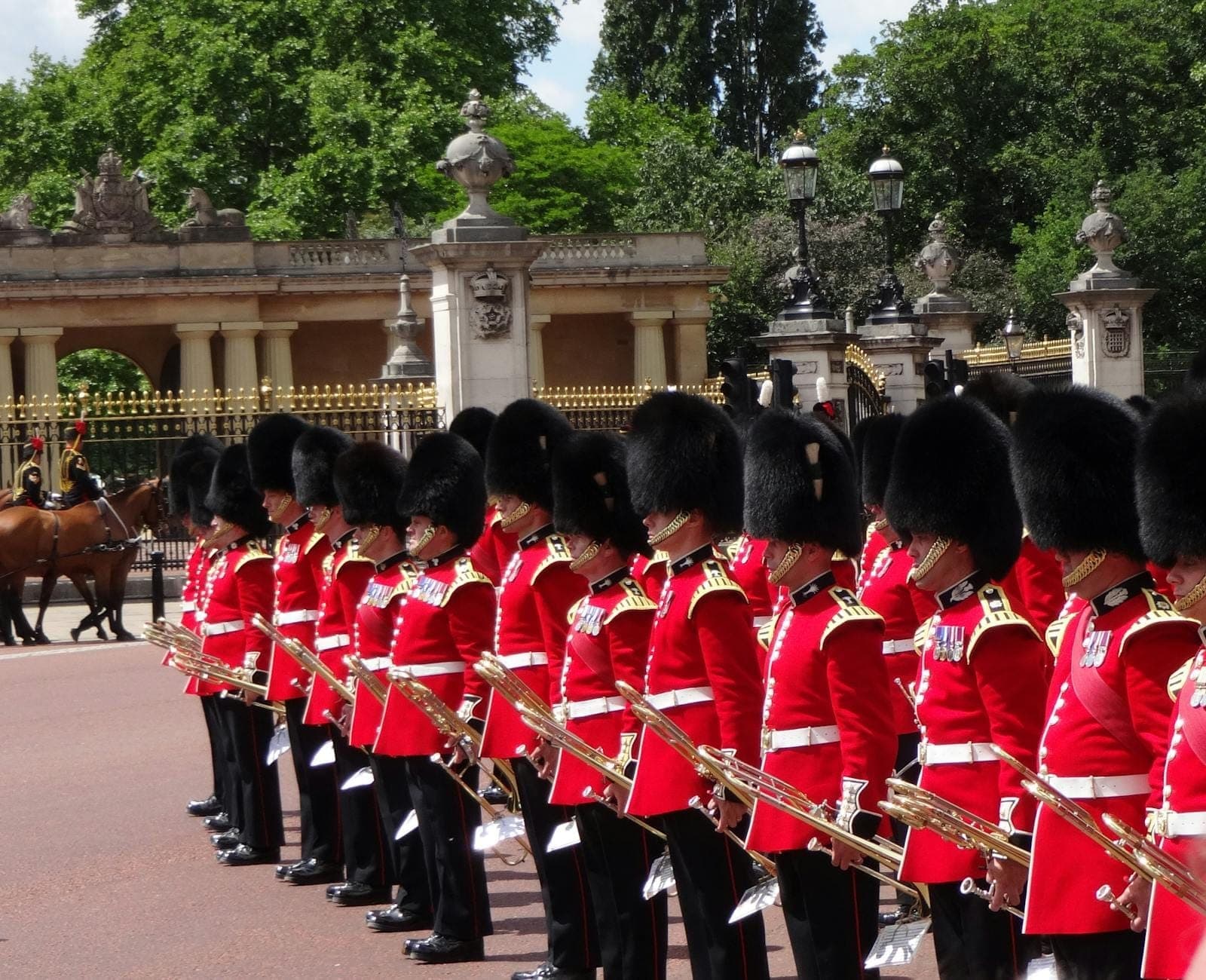 A line of royal guards in red uniforms and bearskin hats stand in formation outside Buckingham Palace, with ornate gates and greenery in the background.