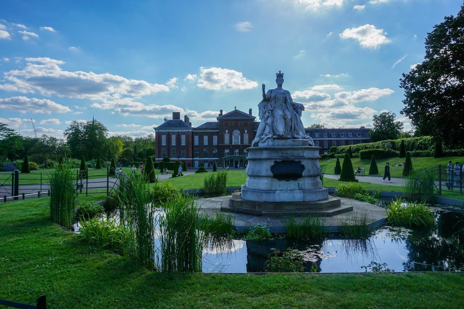 Statue of Queen Victoria in front of Kensington Palace with formal gardens and a reflecting pond under a sunny sky.