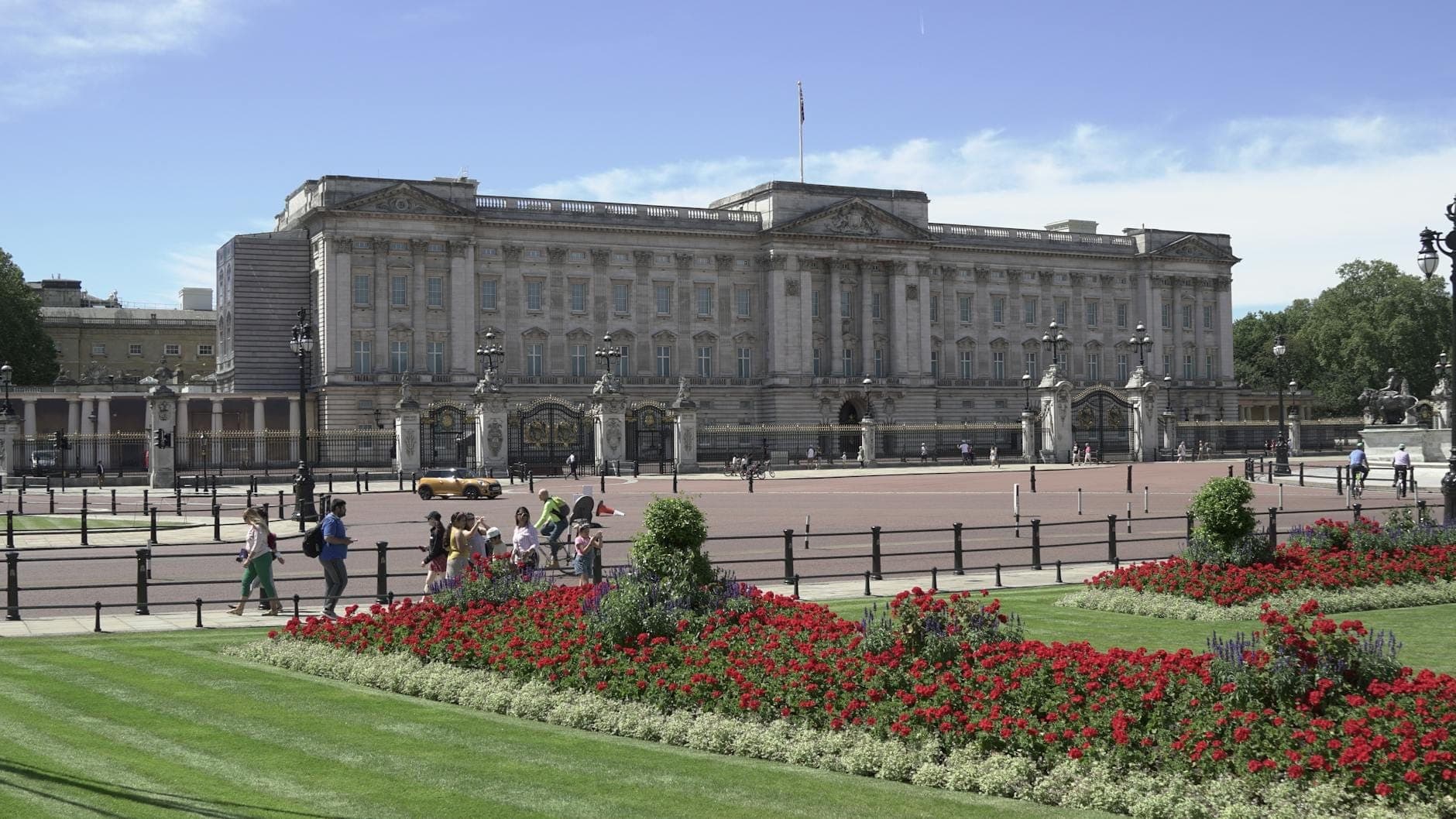 Wide view of Buckingham Palace with its grand facade, front courtyard, ornate gates, and colorful flower beds on a sunny day.