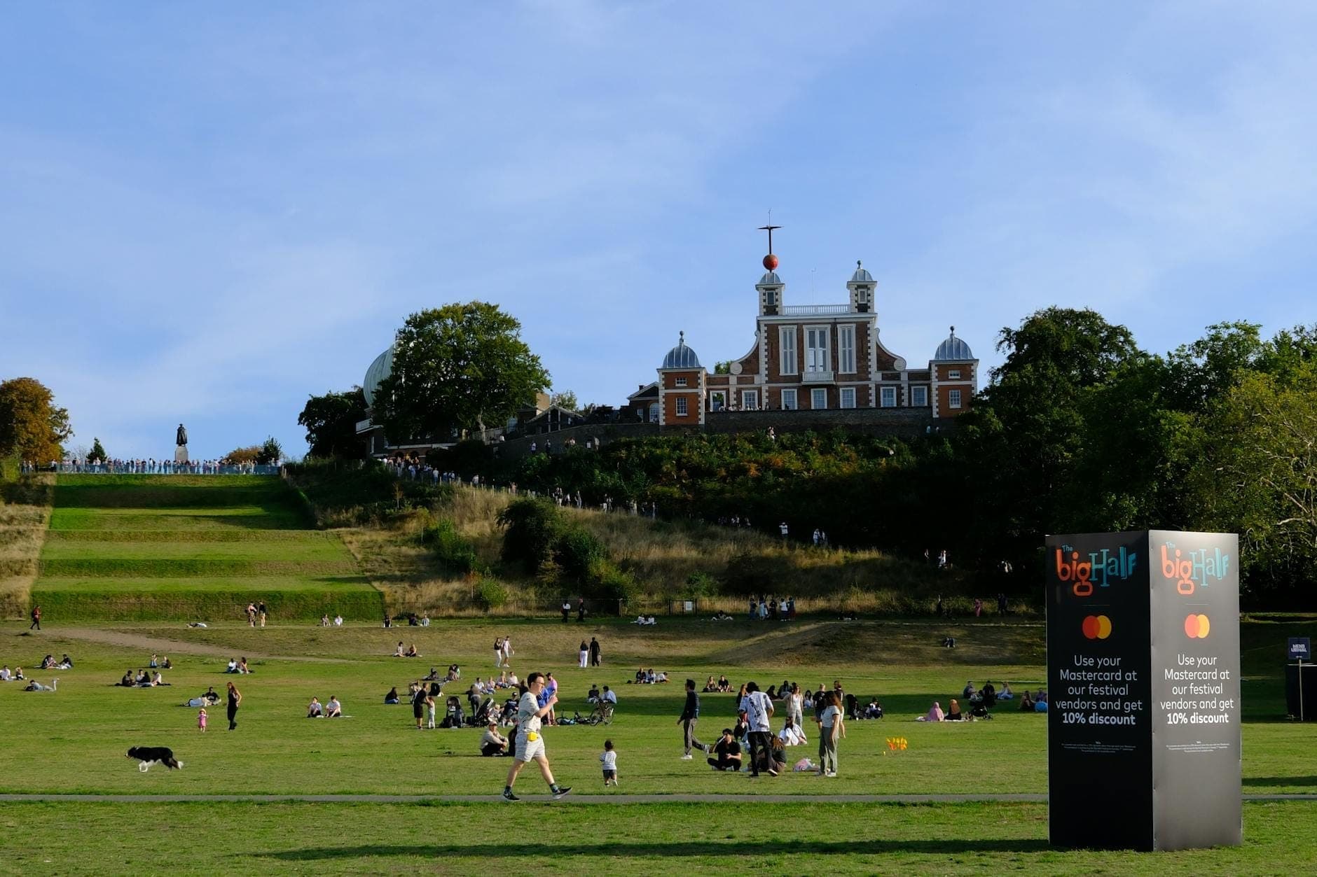 Menschen entspannen auf dem Grashang unterhalb des Royal Observatory Greenwich; der rote Time Ball hebt sich deutlich vom blauen Himmel ab, umgeben von grünen Bäumen.