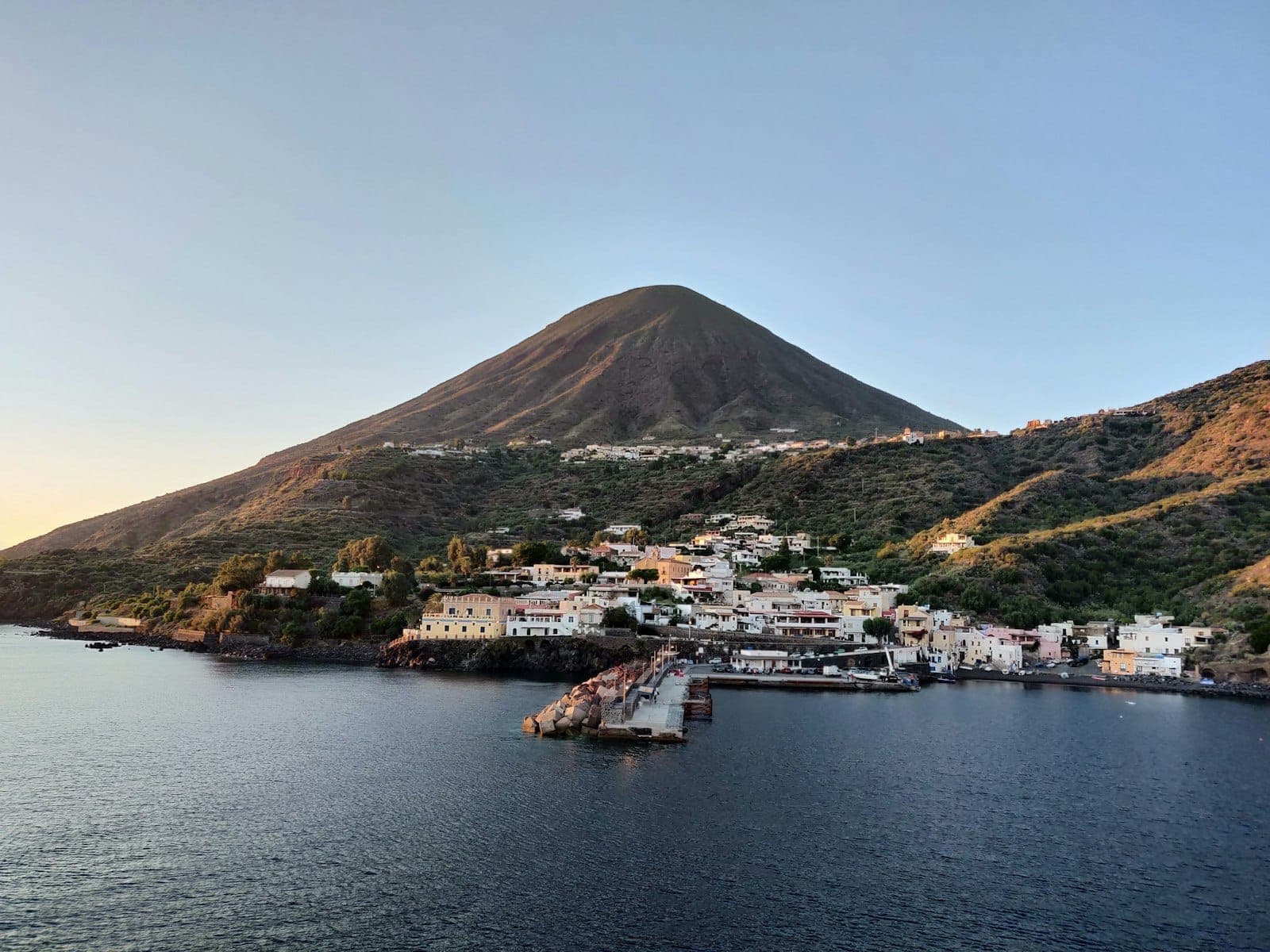 Vista panoramica del villaggio costiero dell'Isola di Salina e dei lussureggianti pendii vulcanici verdi sotto un cielo azzurro limpido al tramonto.