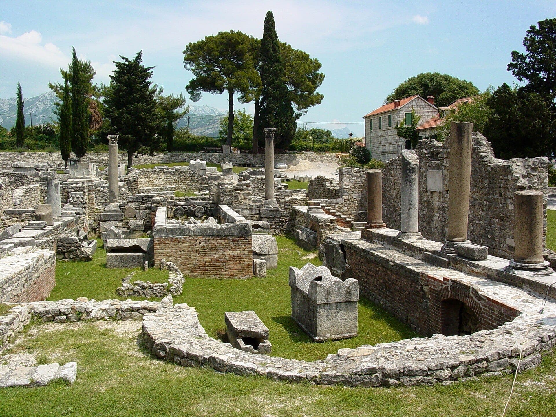 Rovine in pietra e colonne in piedi delle Rovine Romane di Salona, circondate da cipressi e montagne lontane sotto un cielo azzurro brillante.
