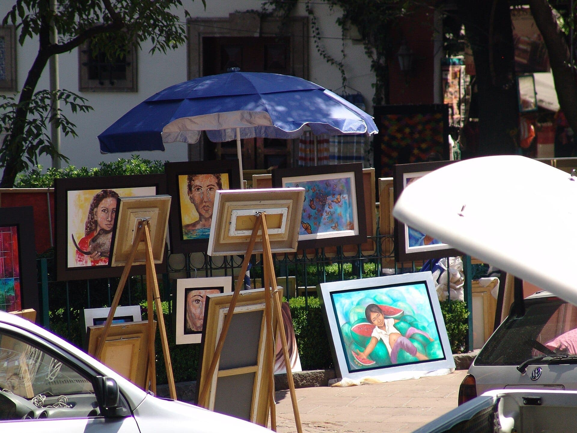 ตลาดงานศิลปะกลางแจ้งที่ Bazar del Sábado ใน San Ángel มีภาพวาดสีสันสดใสบนขาตั้งภาพใต้ร่มสีน้ำเงิน ล้อมรอบด้วยรถยนต์และต้นไม้สีเขียว