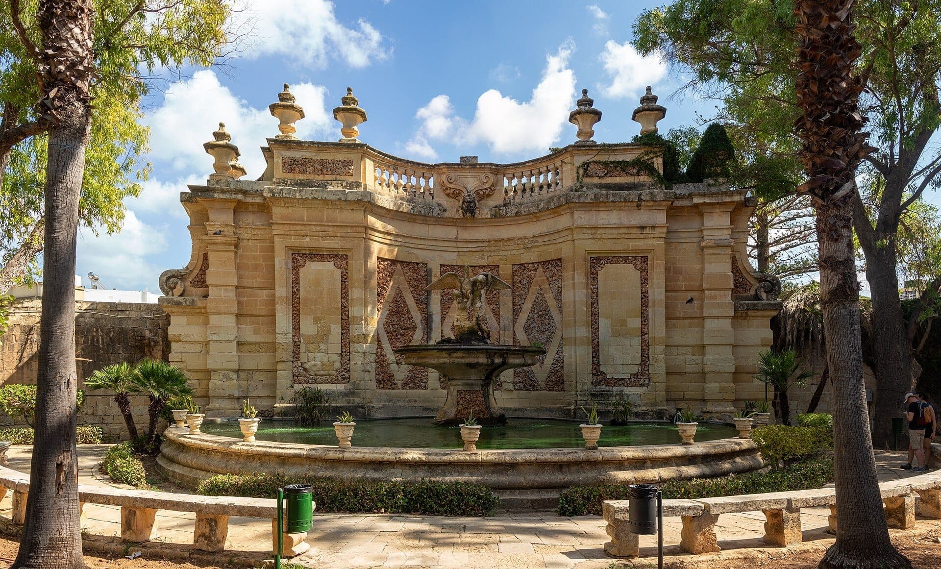 Zentraler Brunnen mit einer geflügelten Statue, umgeben von historischer Sandsteinarchitektur und üppigem Grün in den San Anton Gardens bei hellem Tageslicht.