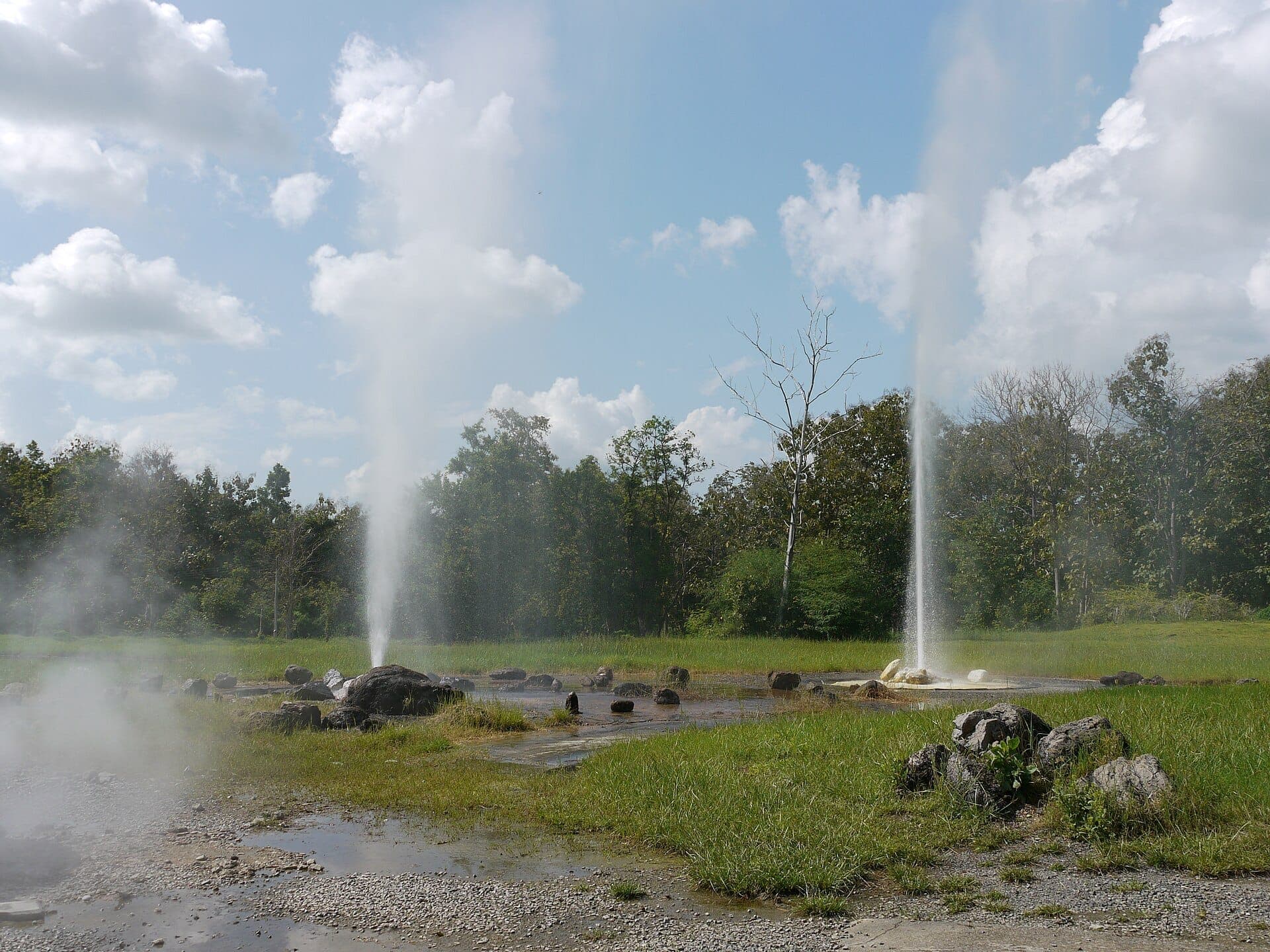 Natürliche Geysire in den San Kamphaeng Hot Springs brechen vor einem Hintergrund aus grünen Bäumen, Felsen und klarem blauem Himmel bei Tageslicht aus.