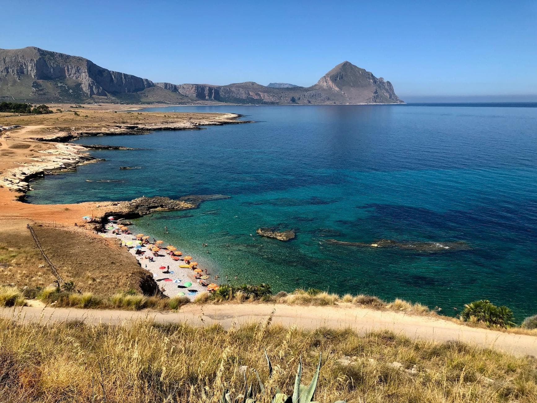 Pemandangan indah Pantai San Vito Lo Capo dengan air pirus jernih, pasir keemasan, payung pantai warna-warni, dan tebing Monte Monaco yang dramatis di latar belakang.