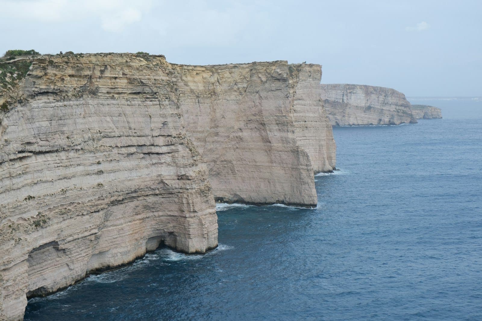 Die steilen Kalksteinfelsen der Sanap Cliffs ragen dramatisch über das tiefblaue Mittelmeer an der unberührten Südküste von Gozo auf, unter einem leicht bewölkten Himmel.