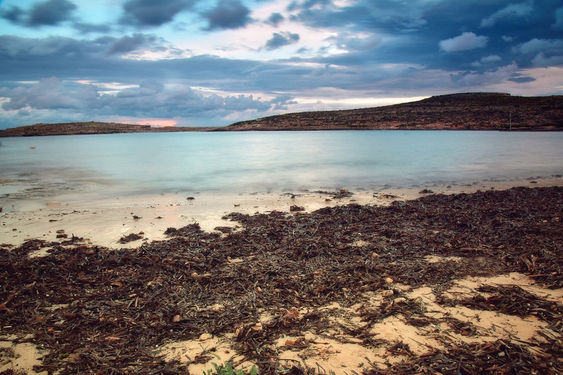 Küste der Santa Marija Bay mit sandigem, von Algen bedecktem Strand, ruhigem türkisfarbenem Wasser und felsigen Hügeln unter einem bewölkten Himmel in der Abenddämmerung.
