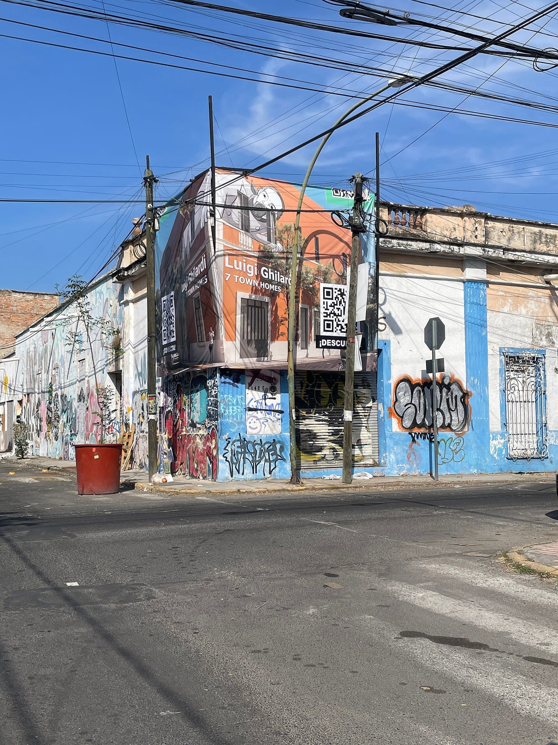 มุมถนนใน Santa Teresita, Guadalajara มีกราฟฟิตีสีสันสดใส ตึกเก่า ป้ายโฆษณาขนาดใหญ่ และสายไฟที่พาดตัดกันใต้ท้องฟ้าสีฟ้าใส